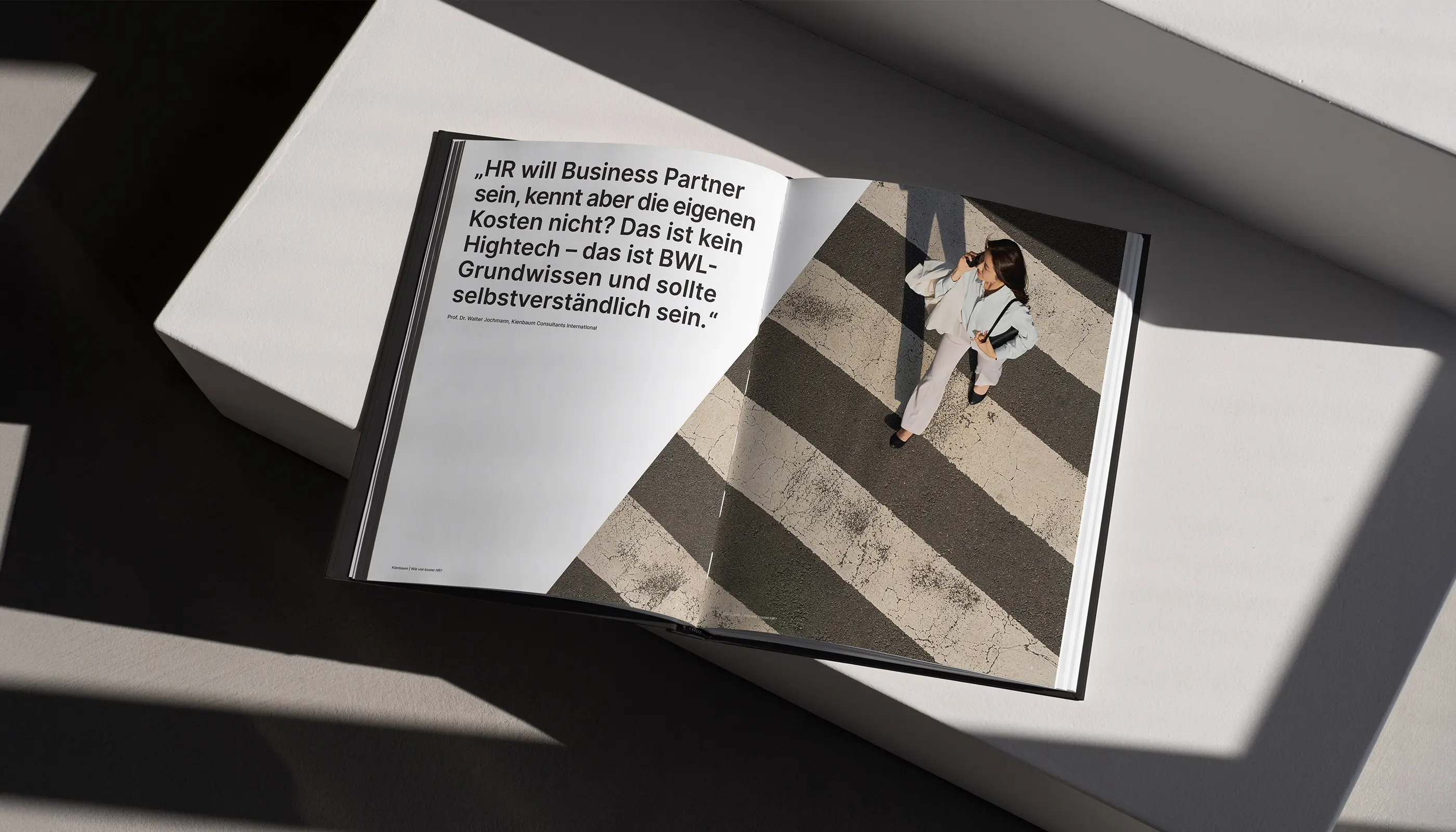 Open book showing a quote in German on the left page and a top-down photo of a woman walking on a pedestrian crossing while talking on her phone on the right page.