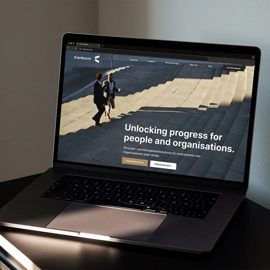 Laptop on dark surface displaying Kienbaum website with two men walking on outdoor steps and headline 'Unlocking progress for people and organisations.'
