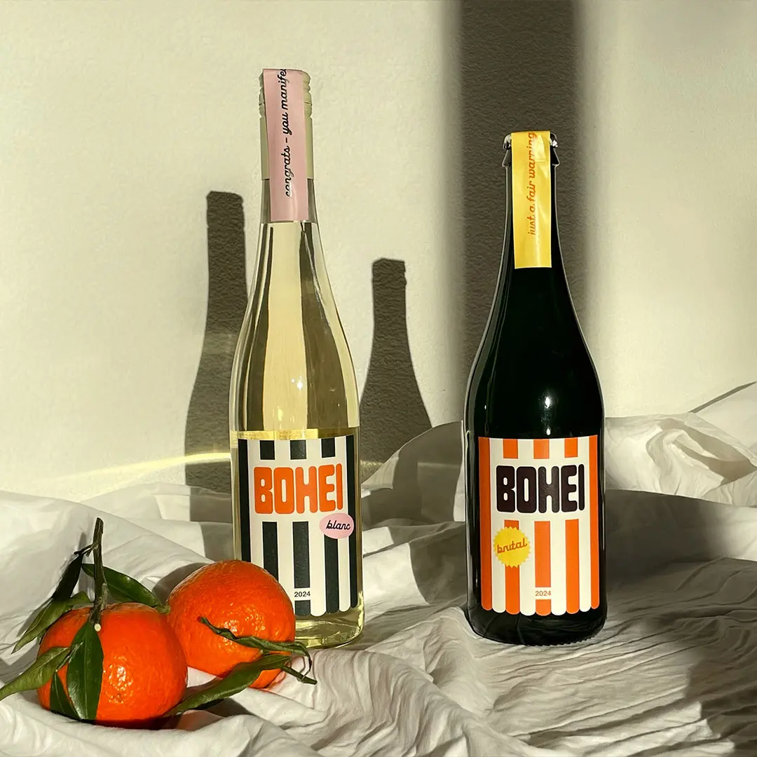 Two bottles of Bohei wine, one white with black and white stripes labeled 'blanc' and one darker with orange and white stripes labeled 'brutal', placed on white fabric with three fresh oranges beside them.