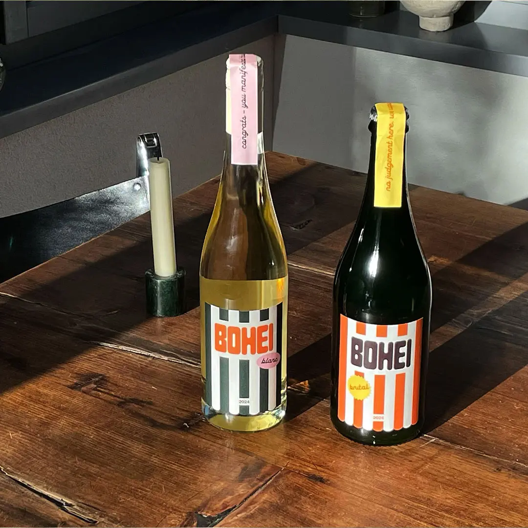 Two bottles of Bohei wine, one white with pink label and one dark with orange label, on a wooden table with a candle holder nearby.