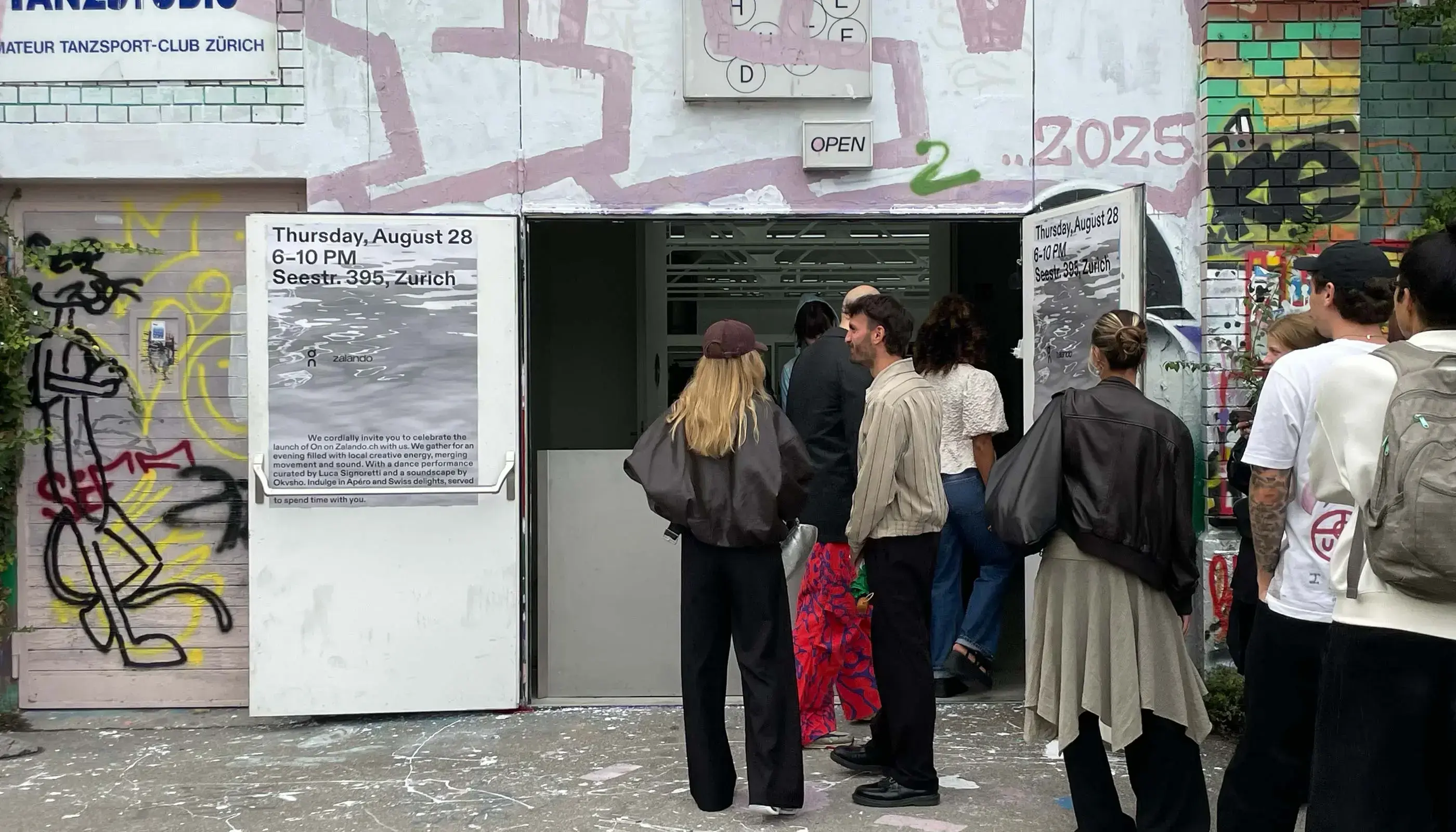 People standing outside a graffiti-covered building with open doors displaying signs for an event on Thursday, August 28, 6-10 PM at Seestr. 395, Zurich.