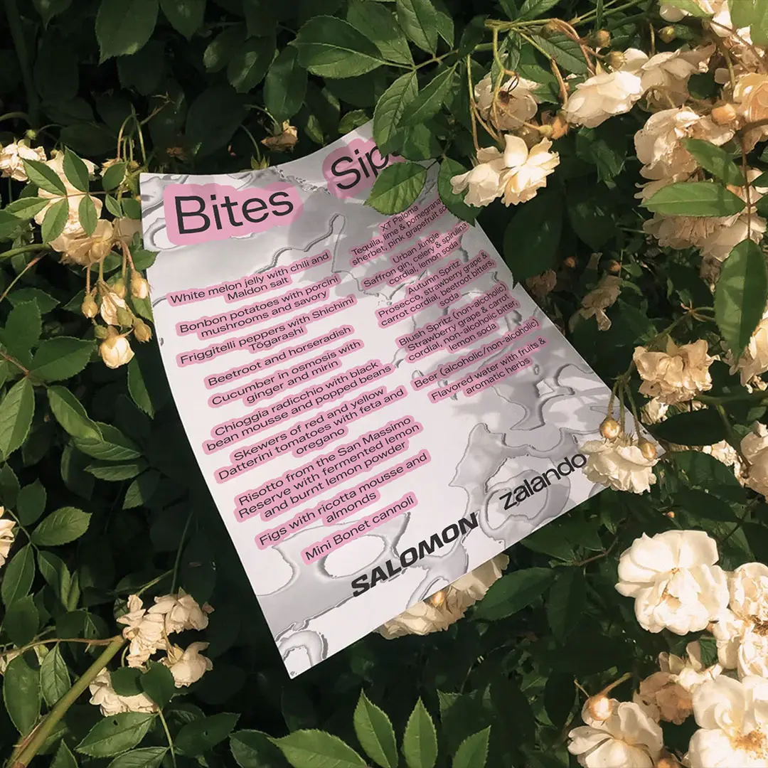 Menu card titled 'Bites' and 'Sip' with food and drink items, placed among green leaves and white flowers.