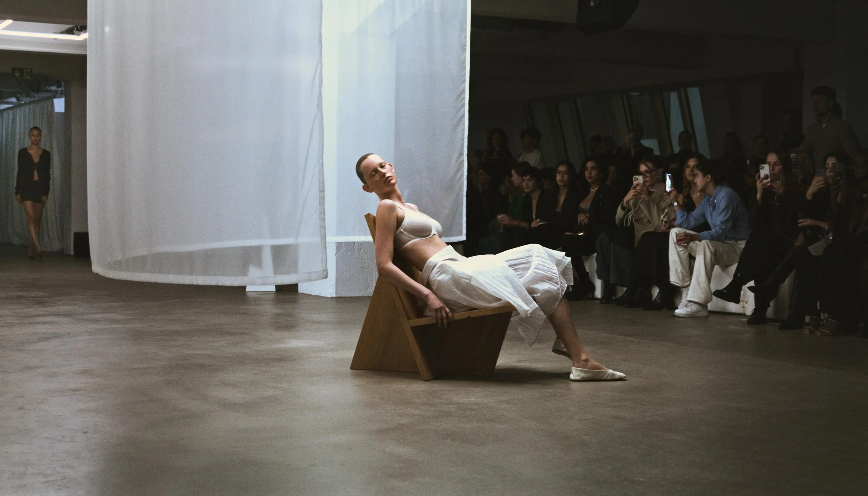 Fashion model in white skirt and bra sitting sideways on a modern wooden chair during a runway show with an audience watching and another model walking in the background.